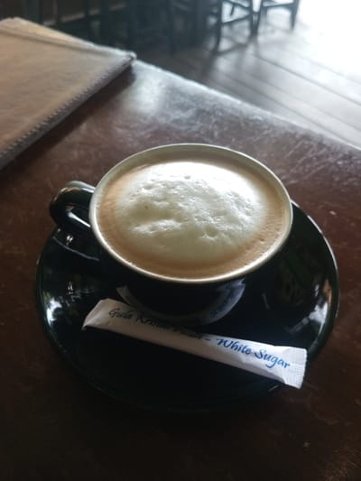 A cup of frothy coffee sits atop a black saucer on a dark wooden table. A white packet of sugar rests beside the cup. The coffee has a light brown hue beneath the thick layer of foam. The table surface is slightly glossy, reflecting some light. In the background, blurred shapes suggest chairs and a window. A menu or notebook is partially visible in the top left corner.