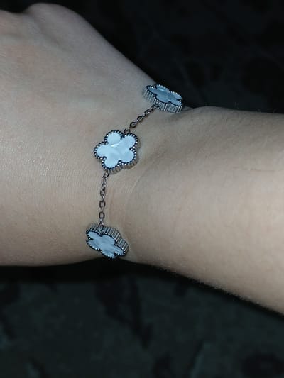 A close-up, overhead shot shows a white mother-of-pearl clover bracelet with silver trim on a human arm. The bracelet has three clover charms connected by a delicate silver chain. The bracelet is positioned diagonally across the forearm, with the charms facing upwards and slightly to the right. The skin appears fair, with some visible texture and fine hairs. The background is dark and out of focus, with a hint of a textured surface, possibly a dark fabric or carpet. The lighting is dim, casting shadows and highlighting the reflective surfaces of the bracelet. There are no discernible landmarks or elements within the image that would allow for identification of a specific location in Korenovsk, Russia.