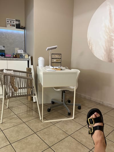 In Saint-Galmier, France, an interior scene depicts a small desk with a white chair and a desk lamp. On the desk, an earring stand displays multiple pairs of earrings. A white trash can is positioned near the desk. To the left, a wicker chair is visible, partially covered by a light-colored cushion and a tan-colored throw. A framed picture is on the wall above a shelf containing various items. The floor is tiled in a neutral color. In the foreground, a person's foot is visible wearing a black sandal with gold buckles.