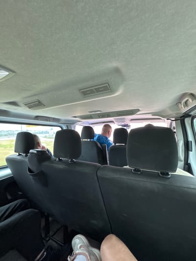 The image captures the interior of a vehicle, likely a van or shuttle, viewed from a passenger seat looking towards the front. The light-colored ceiling, running along the top of the frame, appears textured and somewhat dusty or stained, with rectangular air vents visible along its length.

In the middle and lower sections of the frame, several rows of black fabric seats and headrests are visible, indicating a multi-passenger vehicle. The back of at least three headrests are clearly seen. Further forward, a person wearing a blue short-sleeved shirt is seated, facing away from the camera, their head and upper back partially visible. In the immediate foreground, towards the bottom right, a person's arm and a foot wearing a white and light pink athletic shoe are visible, suggesting the vantage point is from a seat behind them or beside them, looking forward.

Through the vehicle's windows, seen on the far right, a blurry green landscape is visible, indicating movement or being stopped in an outdoor environment during the daytime. The lighting is bright and natural, suggesting clear or overcast weather conditions. The scene depicts people being transported in the vehicle. No discernible text is visible anywhere in the image. Given the context of Kelsterbach, Germany, the setting could be near an airport or a travel hub.