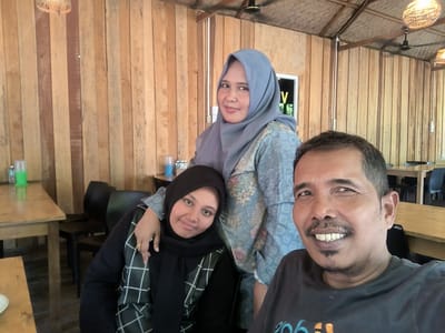 The image shows three people posing indoors for a photo. The location appears to be a restaurant or dining area with wooden walls and ceiling. The lighting is somewhat bright, possibly due to natural light.
From left to right: a woman wearing a black headscarf and plaid pattern shirt is smiling at the camera. The woman in the middle is wearing a blue headscarf and a floral pattern top. She is smiling at the camera. On the right, a man is smiling directly at the camera, wearing a dark gray shirt. Tables and chairs are visible in the background, further supporting the idea of a restaurant setting. Other objects are present in the background, including drink containers on a table. The image captures an interpersonal moment. The location is the city of Santan, Indonesia.