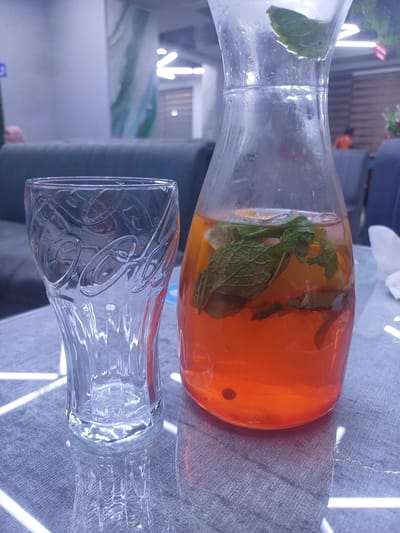 An empty Coca-Cola branded glass stands to the left of a glass carafe filled with an orange beverage, infused with mint leaves and citrus slices. Both items are placed on a textured grey tablecloth with a white geometric pattern. The background is blurred, showing a seating area with dark sofas and a woman in an orange shirt in the distance, suggesting an indoor establishment. The scene appears to be a restaurant or cafe, possibly in Shahand, Uzbekistan.
