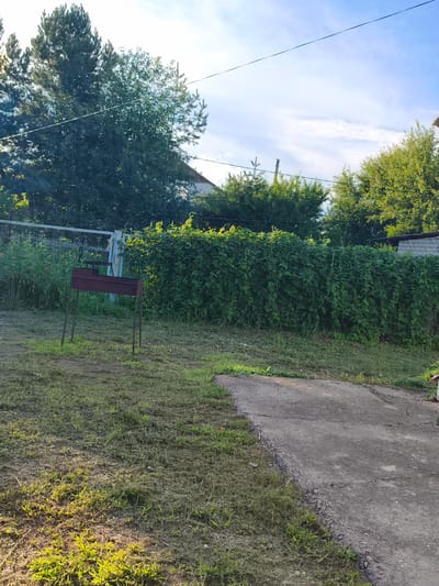 The image shows a backyard scene in Konstantinovskaya, Russia. A maroon metal grill is standing on a mown lawn. Behind the grill, a dense green hedge runs horizontally across the frame. Beyond the hedge, there are trees and a building with a brick wall. The sky is visible above, with some clouds. A concrete path is present in the foreground.