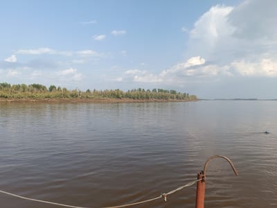 The image depicts a wide, calm river with a tree-lined bank in the distance. The sky is partly cloudy, with a large cloud formation on the right side of the frame. In the foreground, a reddish-brown metal post with a curved hook is visible, along with a rope extending across the bottom of the image. A small blue object floats on the surface of the water to the right. The overall scene suggests a tranquil, natural environment, possibly a waterfront area in or near Alexandrovskoe, Russia.