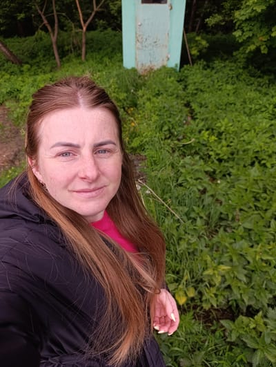 The image shows a woman in a black jacket and pink shirt, with long reddish-brown hair, fair skin, and visible freckles. She is smiling slightly and looking directly at the camera. The background consists of lush green vegetation. A small, weathered turquoise structure resembling an outhouse is visible in the distance behind her. The scene appears to be outdoors, possibly a park or wooded area. The location is stated to be in the city of Chernogolovka, Russia.