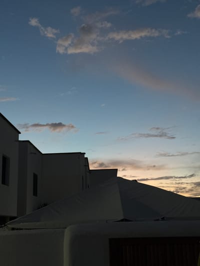 The image, correctly oriented, captures a tranquil sky scene above buildings in Yaiza, Spain, indicative of either sunrise or sunset. The sky transitions from a clear, deep blue on the left to warm, soft peach and orange tones on the right, illuminated by the low angle of the sun. Scattered, wispy clouds, varying from light grey against the blue to subtly lit by the warm light, are dispersed across the sky.

In the foreground and along the right edge, the stark silhouettes of angular, white buildings, characteristic of local architecture with flat roofs, are visible. These structures appear dark due to backlighting. A large white canvas awning or canopy partially obscures the upper right, and a dark, rectangular window or shutter is discernible within one of the building facades. No people or activities are depicted, and no text is visible. The scene evokes a sense of peacefulness and natural beauty.