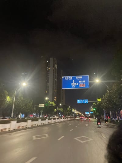 This image captures a wide, multi-lane urban street scene at night in Dali City, Yunnan Province, China. The perspective is from within a vehicle, looking forward.

The precise location is identified by a prominent blue overhead road sign, which indicates directions for "云岭大道" (Yunling Avenue), "富海路" (Fuhai Road), and "巍山路" (Weishan Road), also designated as National Highway G320. The sign shows arrows directing straight ahead/left for Yunling Avenue, and right for Fuhai Road and Weishan Road/G320, along with a U-turn symbol.

The asphalt road surface is wet, reflecting the numerous bright streetlights that line both sides of the street, producing significant lens flares. White dashed and solid lane markings are visible, along with several square white markings embedded in the pavement. A long, white, segmented concrete median barrier topped with dark vegetation separates the traffic lanes.

Multiple vehicles with illuminated taillights are observed in the distance, traveling away from the camera. On the far right lane, two individuals are seen operating scooters or electric bicycles, also moving away. Tall, multi-story buildings, some with illuminated windows, are visible on the left side of the street, set back from the road. Additional streetlights and trees line the thoroughfare. The sky above is dark and overcast. The lower right foreground shows a dark, indistinct mass, likely part of the vehicle from which the photo was taken.