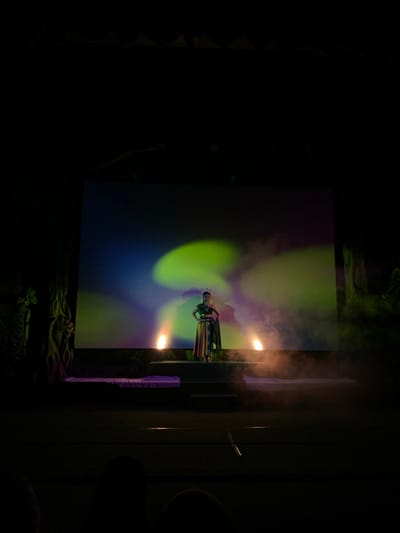 The image shows a stage performance in a dark theater setting. A single female performer is standing on a raised platform, facing the audience. She wears a colorful, patterned outfit with gold embellishments.  The stage is lit by two spotlights, creating a hazy, atmospheric effect.  The background features a large screen displaying a dynamic, colorful abstract image of a green, blue, and purple aurora-like pattern.  The performance is taking place at the Jatim Park 3, Batu Secret Zoo, in Batu, Indonesia, located at the coordinates -7.884, 112.524.  The setting evokes a mystical and dreamlike atmosphere.