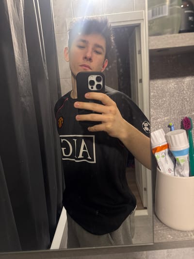 The image depicts a mirror selfie taken in a bathroom in Yanino-1, Russia. A young male, with short dark hair, is the sole subject, positioned centrally in the reflection. He is wearing a black Manchester United football jersey featuring the "AIG" logo in white on the front, and light grey sweatpants. His right hand holds a black smartphone, featuring a triple-lens camera array, positioned to capture his reflection. To the left, a dark grey shower curtain is visible. To the right, a light-colored cup contains two tubes of toothpaste, one with orange accents and one with light blue accents, along with two toothbrushes, one green and one purple/pink. A textured grey wall or tiling is visible in the background, above which a dark bottle rests on a shelf. A white door frame is partially visible in the reflection behind the subject. Overhead lighting creates a bright glare in the upper central portion of the image.