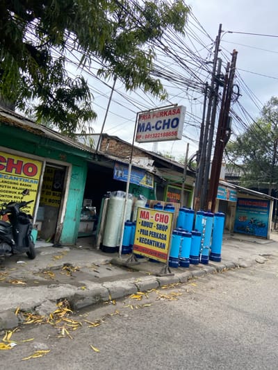 The scene is a street in Kecamatan Margahayu, Indonesia, located at latitude -6.961, longitude 107.579. There is a small shop on the left side of the image with a sign that says "CHO". The shop appears to be selling beauty products. To the right of the shop is a building with a large sign that says "PRIA PERKASA MA-CHO HP: 081322188656" in red and white letters on a blue background. In front of the building is a row of blue cylindrical objects, possibly water filters, and another sign that says "MA-CHO ADULT - SHOP PRIA PERKASA KECANTIKAN HP: 081322188656". There are many power lines overhead. There is a motorcycle parked on the left side of the image. There are dry leaves scattered on the sidewalk. The street appears to be paved.