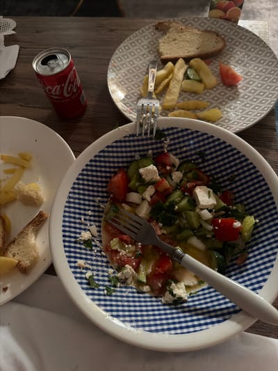 A still life photograph captures a casual meal setting on a wooden table. The scene features a red Coca-Cola can, a plate with bread, fries, and a slice of tomato, and a bowl of salad with feta cheese, tomatoes, and cucumbers. A fork rests in the salad bowl, and another fork is partially visible on the plate with fries. The overall impression is one of a simple, possibly home-cooked meal. The location of Dromolaxia - Meneou Municipality, Cyprus, suggests a Mediterranean context for the meal, which is consistent with the ingredients observed.