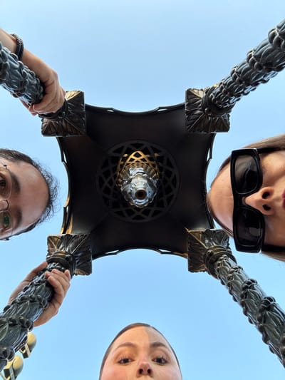A low-angle shot features three people surrounding a tall, ornate lamppost. The woman at the top wears sunglasses, with the lamp directly below. To the right, another woman's face is partially visible. Below the lamp, a man holds the post, his face also partially visible. The light blue sky is visible in the background. The lamp itself is dark in color with decorative patterns.