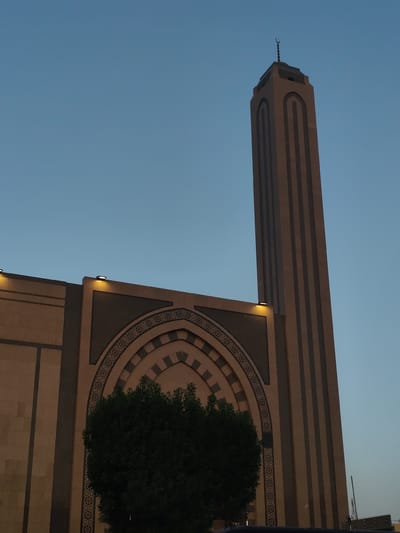 The image shows a tall minaret and part of a building, likely a mosque, against a blue sky. The minaret is slender and rectangular with vertical stripes along its length. At its top, there is a crescent moon symbol, a common feature of mosques. The building has an arched entrance or facade decorated with geometric patterns. There is a dark green bush or small tree in front of the building, partially obscuring the lower part of the arch. Small lights are visible above the arch. The architecture suggests this is a mosque in Makkah Al Mukarramah, Saudi Arabia, but without further context, it is impossible to identify the specific mosque. The lighting indicates it is dusk or dawn.