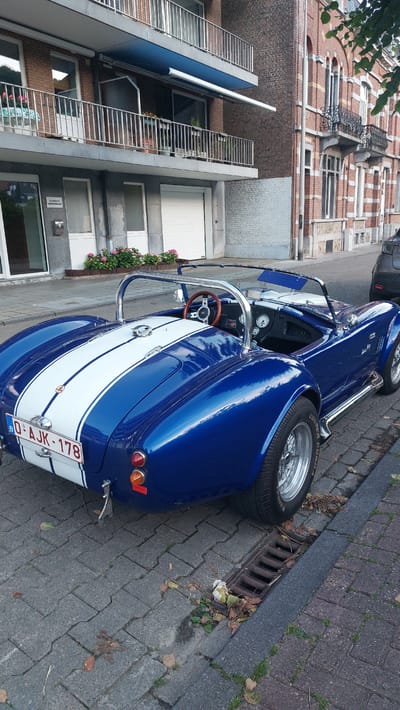 A blue and white AC Cobra convertible sports car is parked on a brick-paved street in Namur, Belgium. The car has a chrome roll bar, a wooden steering wheel, and a license plate that reads "0-AJK-178". The car is facing away from the camera, and its rear is visible. The car is parked next to a dark gray modern SUV.

In the background, there are several buildings. The building directly behind the car is a multi-story brick building with balconies and windows. To the left of this building is a lighter-colored building with a garage door and windows. The street is lined with trees and foliage. The weather appears to be overcast.