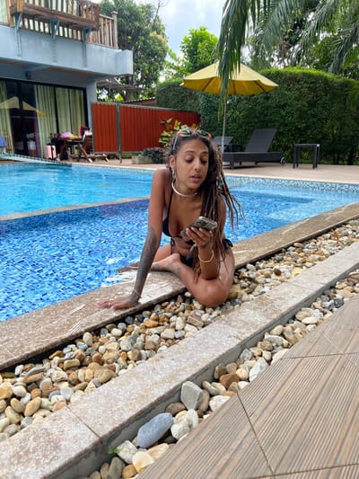 In Ban Tha Muang, Thailand, a young adult female is depicted sitting on the edge of a swimming pool. She has dreadlocks, multiple facial piercings, and tattoos on her left arm and right thigh. She wears a black bikini, a silver choker, and sunglasses pushed onto her head. Her right hand holds a light-colored smartphone, which she is observing, while her left hand rests on the pool coping. Her right leg is bent, and her left leg extends partially into the blue-tiled pool.
The immediate foreground features a section of brown wooden decking and a narrow trench filled with an assortment of small, smooth pebbles. The pool's edge consists of a light-colored stone coping. In the background, a multi-story building with large glass doors and an upper-level balcony is visible to the left of the pool. Outdoor furniture, including a wooden table with chairs and a dark suitcase, are positioned near this building. Further back, a dark-colored lounge chair sits under a yellow market umbrella, adjacent to a dense green hedge. Lush green foliage and trees surround the entire pool area, including palm fronds partially obscuring the sky. The scene is illuminated by natural daylight.