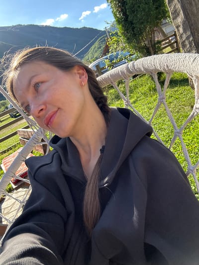 A young woman with braided brown hair, wearing a black hooded jacket and a silver necklace, sits in a white rope hammock. She has blue eyes and is looking to her left, with a slight smile on her face and gloss on her lips. She is outdoors, with lush green grass and trees surrounding her. In the background, there are mountains under a clear blue sky with some clouds. The scene appears to be in a rural or mountainous area, possibly a backyard or a recreational space. The lighting suggests it is daytime, with the sun casting shadows.