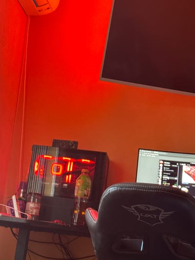 The image captures an indoor computer setup bathed in a warm, reddish-orange light. The walls of the room are a striking orange color, contributing to the dominant hue.
On the left side of the frame, a large, dark monitor stands, its screen reflecting some of the ambient light. Below it, a white power strip or similar electronic device rests on the floor, with a white cable running along the base of the wall.
Towards the right, a black gaming chair is prominently featured. Its headrest displays a distinct logo featuring the stylized letters "CXT" in white. Partially visible behind and to the right of the chair is a second computer monitor, which is active and displays a graphical user interface, resembling video editing software or a streaming application with multiple tracks and visual elements.
Below the second monitor and slightly to the left of the chair, a desktop computer case is visible. It is internally lit with vibrant red LED lights, creating a glow that emanates from its vents and internal components. On top of or immediately next to the computer case, two bottles are placed: a tall green bottle with a yellow cap (possibly olive oil or a beverage) and another darker bottle. Various cables are visible in the background, connecting the devices.
The overall scene suggests a personal workspace or gaming station, characterized by its distinctive lighting and functional setup. No people are visible, and the time of day is indeterminate due to the artificial lighting.