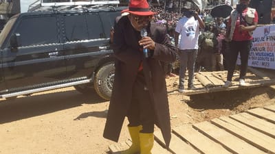 A man with a red hat, glasses, and a long black coat addresses a crowd with a microphone while standing on a makeshift wooden platform. He wears bright yellow boots. Behind him, a black SUV with a roof rack is parked. The crowd is visible in the background, with a banner reading "Bureau de Fin" and other text. The ground appears to be dirt or sand.