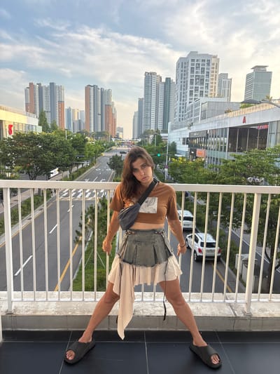 The image depicts a young woman posing for a photo on a balcony overlooking a cityscape. The woman is wearing a brown long-sleeved shirt, a denim skirt with a ruffled hem, and dark slides. She has a dark crossbody bag slung over her shoulder. Her hair is long and brown, and she is looking directly at the camera with a slight smile.

The setting is an urban environment with numerous tall buildings visible in the background. The buildings appear to be apartment complexes and commercial establishments. A road with multiple lanes and yellow lane markings is visible below the balcony, with cars driving on it. Green trees and foliage are interspersed throughout the urban landscape. The sky is partly cloudy, suggesting it is daytime, possibly late afternoon or early evening.

The balcony itself has a white railing and a concrete floor. The woman's pose and expression suggest she is relaxed and confident. The overall impression is a casual snapshot taken during a warm day in a modern city.

Visible text includes "iGo" on a building in the lower right corner. Other text on buildings in the background is too small to be legible.