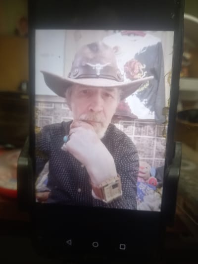 A portrait of a man wearing a cowboy hat and a dark patterned shirt is displayed on a smartphone screen, which is likely in Maloye Lugovoye, Russia. The man has a white beard and is resting his chin on his hand. He is wearing a ring with a turquoise stone on his right index finger and a watch on his left wrist. In the background, a t-shirt with a graphic design is hanging, and a stuffed toy is visible. The overall setting appears to be indoors, possibly a home environment, with a tiled wall behind the man. The smartphone itself is positioned on a dark surface, possibly a table, with navigation buttons at the bottom of the screen.
