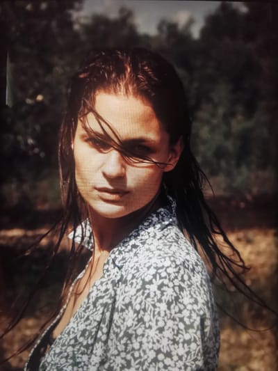 A close-up, head-and-shoulders shot shows a woman with dark, wet hair blowing across her face as she looks directly at the camera. She is wearing a light-colored shirt with a dark floral pattern. Her expression is serious. The background is blurred, consisting of dark foliage and a lighter, earthy-toned area, possibly suggesting an outdoor, natural setting. There are no identifiable landmarks or structures that would indicate the city of Serov, Russia.