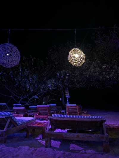 The image shows an outdoor seating area at night in Santa Teresa, Costa Rica. There are several wooden benches with blue and white striped cushions arranged in a row. Two wicker globe lights hang above the seating area, illuminating the space with a warm glow. The benches are situated on a sandy beach, casting long shadows on the ground. Lush, leafy trees surround the area, creating a tranquil and secluded atmosphere. The ground is covered in soft, white sand. The scene is quiet and peaceful, offering a glimpse of a relaxing evening in a tropical paradise.