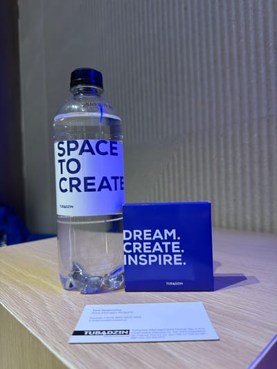 A bottle of water labeled "SPACE TO CREATE TUBADZIN" sits on a light-colored surface next to a blue box that says "DREAM. CREATE. INSPIRE. TUBADZIN." A business card for TUBADZIN is also present. The card contains contact information including Neli Stojanowa Area Manager Bulgaria, and the email address n.stojanowa@tubadzin.pl. Additionally, Tubądzin Management Group Sp.z o.o., codrowice Parcela 11.95-035 Ozorków. Rho, Italy is not explicitly stated in the objects visible.
