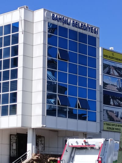 The image shows the exterior of the Bahşılı Belediyesi (Bahşılı Municipality) building in Turkey. The building features a modern white facade with large blue-tinted glass windows. The words "BAHŞILI BELEDİYESİ" are visible in large blue letters on the top of the building. To the right, a yellow sign with black text reads "T.C. BAHŞILI BELEDİYE BAŞKANLIĞI" (Republic of Turkey Bahşılı Municipality Directorate). Below this, there are several framed images or posters showcasing urban development projects, including one that reads "15 TEMMUZ ŞEHİTLER MEYDANI DÜZENLEME YAPIM İŞİ" (15 July Martyrs Square Arrangement Construction Work). In the foreground, part of a white garbage truck with red accents is visible. The sky is clear and blue.