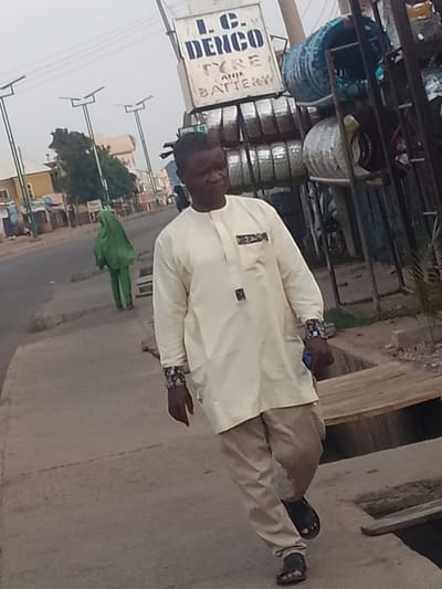 A man in a cream-colored traditional outfit and light-colored pants walks on a sidewalk. He is wearing dark sandals and has a colorful bracelet on his left wrist. In the background, a person in a bright green hijab walks on the street. The scene appears to be a street in Pantame, Nigeria, with a sign for "L.C. DENGO TYRE AND BATTERY" visible. There are also streetlights and what appear to be stacks of tires and other goods for sale at a stall or shop. The sky is overcast.