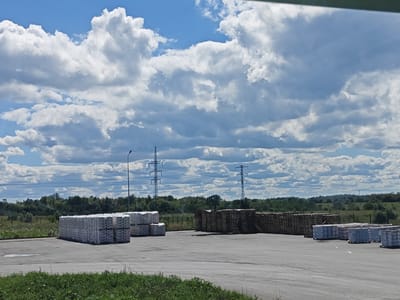An outdoor scene in Chapayevsk, Russia, depicts a large, open lot covered with a grey asphalt surface, likely a storage or industrial area. In the foreground, a strip of green grass borders the lot. The middle ground is dominated by stacks of white bagged materials, possibly cement or building supplies, neatly arranged on pallets. To the right of these stacks, a large collection of wooden pallets is piled high, indicating a distribution or manufacturing hub. In the background, a dense line of green trees forms a natural horizon, beyond which a vast expanse of blue sky is filled with dramatic, white, fluffy clouds. Two tall, metal electricity pylons with visible wires crisscross the sky in the distance, hinting at the presence of power infrastructure. A single streetlamp stands near the left side of the scene, its tall, dark pole stark against the bright sky. The overall impression is one of an active industrial or logistical site under a dynamic sky.