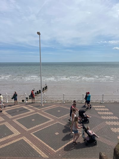 The image captures a section of the Bridlington seaside promenade on a partly cloudy day. A paved walkway, laid with a patterned brick design in shades of brown and beige, runs diagonally across the foreground. Several people are walking along this promenade. To the left, a woman in denim shorts and a dark top walks with a green bag over her shoulder, accompanied by a man in a red life jacket and carrying a similar bag. Further along, two men walk together, one in a blue t-shirt and red hat. In the middle ground, a white railing separates the promenade from the beach and the sea.

The beach itself is a mixture of sand and pebbles, with some wooden groynes extending into the water. The sea is a muted grey-blue with gentle waves. A lone figure stands on one of the groynes.

In the foreground and mid-ground of the promenade, several groups of people are visible. A family with two young children in strollers is walking towards the viewer. One child is seated in a black stroller, while another is in a similar stroller, both dressed in light-colored clothing. Other individuals and groups are also present, walking in various directions. A tall, white lamppost stands prominently on the promenade, near the railing. The overall scene depicts a typical summer day at the seaside in Bridlington, with people enjoying the coastal environment.