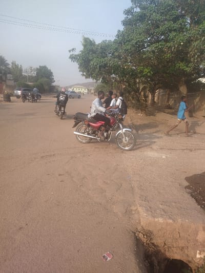 The scene takes place on a dirt road in New Karu, Nigeria, at the coordinates 9 latitude, 7.557 longitude. A man on a red motorcycle is stopped in the middle of the road. There are three other men standing near the motorcycle, two are in white shirts and one is wearing a black shirt. One of the men in a white shirt has a backpack on. In the background, another man in a black shirt is riding a black motorcycle. There are other cars and motorcycles parked on the side of the road. There is a large tree on the right side of the road. A young boy is walking away from the motorcycle towards the tree. There is a large ditch to the right of the road that appears to have eroded away. The scene is sunny and the sky is clear.