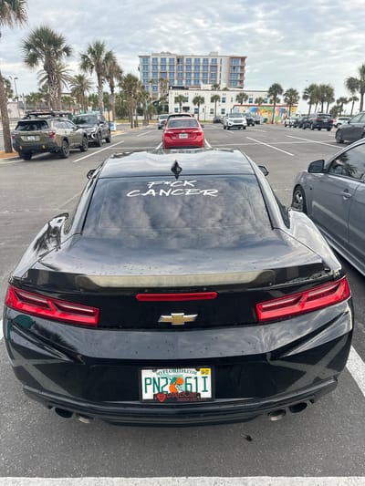 The image shows a black Chevrolet Camaro parked in a parking lot in Jacksonville Beach, Florida, United States of America. The car has a Florida license plate with the number PN2 61T. The phrase "F*ck Cancer" is written in white on the rear window of the vehicle. In the background, there are palm trees, buildings, a colorful mural, and other parked cars. The day appears overcast.