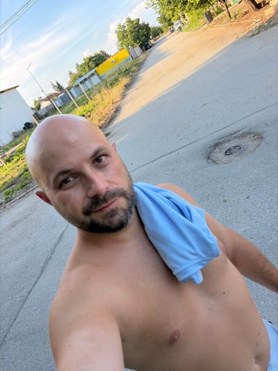 A shirtless, bald man with a beard takes a selfie outdoors. He is wearing light blue shorts and has a light blue shirt draped over his shoulder. The man is standing on an asphalt road with patchy vegetation and several buildings visible in the background. A yellow building with blue trim and a white building are prominent structures. The sky is blue with scattered clouds. The scene appears to be in a rural or suburban area, consistent with a location in Dolni Dabnik, Bulgaria. A drain cover is visible on the road surface in front of the man.