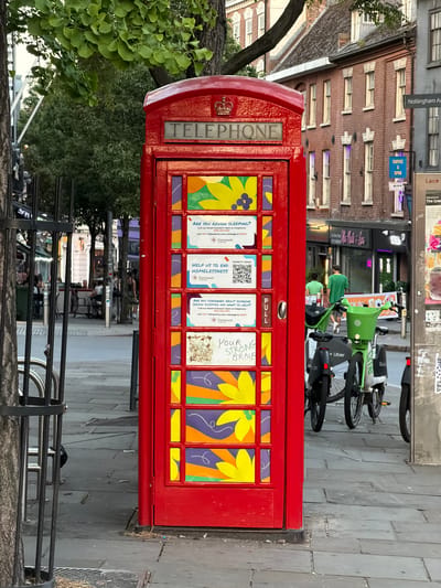 A red telephone booth stands on a paved sidewalk in Nottingham, United Kingdom. The booth's windows are decorated with colorful floral designs and informational signs promoting homelessness assistance programs, including donation QR codes. A green Lime bike and a black Uber bike are parked behind the booth. In the background, a multi-story brick building is visible with signs for "Nottingham Av" and "COFFEE X SPUD". A shop sign reading "After Milk & Soy" is also visible. People can be seen walking and sitting at outdoor tables further down the street. A tree with green leaves partially obscures the booth. A metal railing is in the foreground.