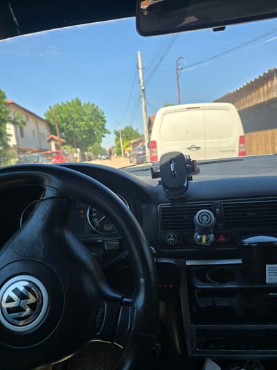 The image shows a view from inside a car looking forward. The steering wheel of a Volkswagen is visible in the foreground. On the dashboard, there is a phone mount holding a smartphone, and an air freshener. The car's dashboard displays the speedometer which reads approximately 40 km/h, and the odometer reads 38352. Outside the car, a white van is directly ahead, followed by houses and trees under a clear blue sky. The location is identified as Skutare, Bulgaria.