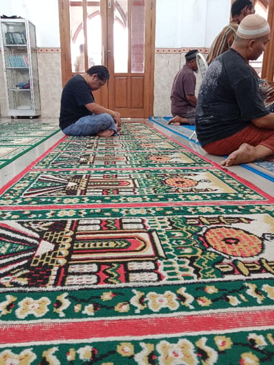 The image shows a mosque in Kecamatan Brangsong, Indonesia (latitude: -6.948, longitude: 110.231). There are three men in the mosque. One man is sitting on a prayer rug, wearing a blue shirt and pants. He is facing towards the camera, looking at the floor. Another man, wearing a white shirt and black pants, is sitting on a prayer rug further down the row. He is facing away from the camera. A third man, wearing a black shirt and pants, is sitting on a prayer rug to the right of the first man. He is facing away from the camera. The floor of the mosque is covered in patterned prayer rugs. The room is lit by natural light. The walls are painted white. The men appear to be praying.