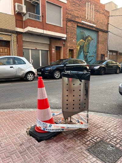 This vertical image depicts a bustling street scene in Picassent, Spain, under bright daylight. In the immediate foreground, an orange and white traffic cone is positioned on a red brick sidewalk, marking a damaged section of pavement. White and blue police tape, bearing the Spanish words "POLICIA LOCAL" (Local Police) and "NO PASAR" (Do Not Enter), is wrapped around the cone and a nearby metal trash bin.

The tall, stainless steel trash bin, perforated with square holes, stands prominently on the sidewalk, containing a black plastic bag. Behind it, on the asphalt street, several cars are parked. A silver compact car is visible at the bottom left, followed by a black sedan, and further down, a black SUV-like vehicle with a partially visible license plate reading "8532 MPJ".

The background features multi-story brick buildings lining the street. One building is adorned with a large, vibrant mural depicting avian figures in shades of green and blue, resembling street art on a rolled-down shutter. Other building details include various windows, some with closed grey roller shutters, brown wooden doors, and external air conditioning units. Visible electrical wires run along the building facades. On the sidewalk, a metal manhole cover is visible, inscribed with "AGUA POTABLE" (Potable Water) and numbers "00" and "300". The scene suggests a temporary restriction or hazard on the sidewalk.