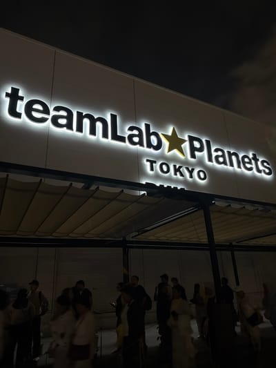 The image shows the exterior of the teamLab Planets Tokyo digital art museum located in Koto, Japan at night. A large, illuminated sign above the entrance reads "teamLab Planets TOKYO". The sign has bright white lettering with a yellow star icon between "teamLab" and "Planets". Below the sign, a retractable awning is partially extended, supported by black metal beams. A crowd of people, some blurred due to movement, are visible standing in a queue or milling around outside the entrance. The environment is dimly lit, with the primary illumination coming from the sign and some ambient lighting.