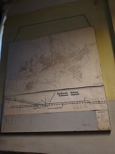 The image shows two large maps mounted on a wall in what appears to be an indoor space with painted walls. The top map is a detailed city plan, depicting streets, buildings, and other urban features. Text above this map reads "TUKUMA PILSETAS PLANS" which translates to "Tukums City Plan" in English. The bottom map is a schematic diagram, possibly of a transportation or utility network, with lines, symbols, and text labels. This map has the title "DZELZCEĻA SHĒMA TUKUMA RAJONĀ" which translates to "Railway Scheme of Tukums District". To the right of the bottom map, there is a legend labeled "Apzīmējumi" which means "Legend" in English, with symbols and corresponding descriptions. The maps are slightly worn and appear to be old. The overall environment suggests a public building, possibly a train station, administrative office, or museum in Tukums, Latvia.