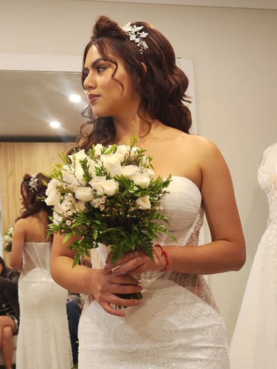 The image shows a woman in a white, strapless, beaded wedding dress. She is holding a bouquet of white flowers and greenery. She has brown, curly hair styled up with a decorative hairpiece. She is looking to her left. In the background, there is a mirror that reflects the woman, another person seated, and another wedding dress. There is a light colored wall and some wood paneling visible in the reflection. It is impossible to confirm this image was taken in Tacna, Peru without additional data.