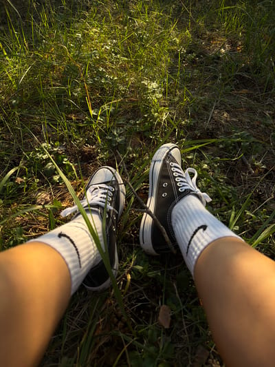 The image shows a pair of feet wearing black high-top Converse sneakers with white laces and white crew socks with a black Nike logo. The feet are positioned in the grass, with sunlight filtering through the blades. The background consists of green grass and dry leaves, suggesting an outdoor environment. The scene is captured from a first-person perspective, looking down at the feet. The location is identified as Udelnaya, Russia.
