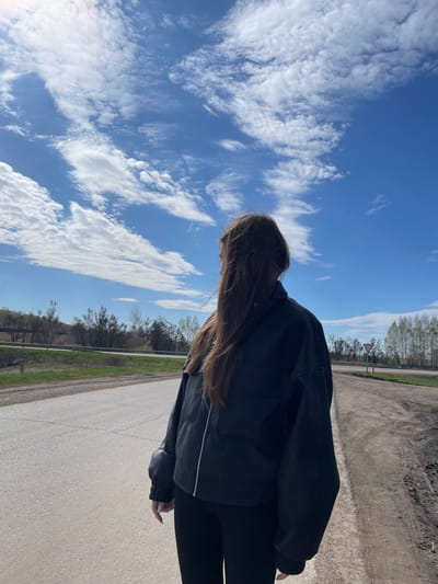 A medium shot shows a person with long brown hair and wearing a black leather jacket. The person stands on a paved road in Ulukulevo, Russia. The road has a strip of green foliage running along the edge and dirt on the other side. Overhead, the sky is blue with scattered white clouds. In the distance, a road sign is visible. The person faces away from the viewer and looks down at the road. They hold something white in their right hand.