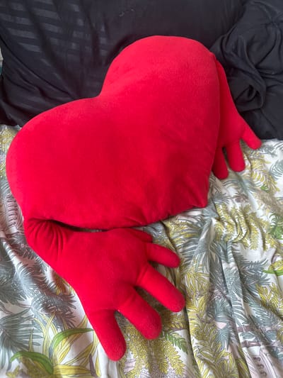 A red, plush pillow with the shape of a heart and attached hands is lying on a bed. The pillow is a vibrant red and has a soft, fuzzy texture. It is positioned diagonally across the frame, with its pointed end towards the lower left and its broader base towards the upper right. Two of the pillow's hands are visible, one extending to the left and the other to the right. These hands are also red and have a similar plush texture. The pillow is resting on a bed with a patterned duvet cover. The duvet features a botanical print with stylized leaves and trees in shades of green, yellow, and brown, set against a white background. To the left of the pillow, a section of a black blanket or sheet is visible, appearing to be made of a slightly shiny material with subtle vertical stripes. The lighting suggests it is daytime, with natural light illuminating the scene. The setting appears to be a bedroom or a living room area where the bed is located. No people are visible in the image, and no text is present.