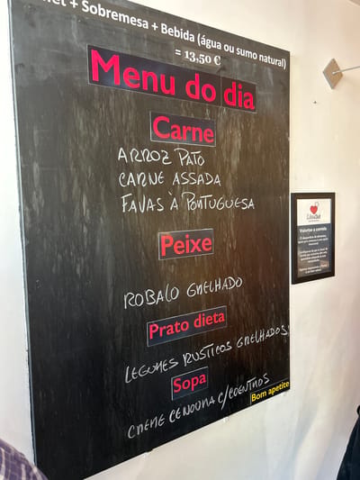 The image shows a chalk menu board in what appears to be a restaurant in Cacém e São Marcos, Portugal. The menu board is black and has handwritten text in white chalk and some red text on black rectangles. The top of the menu states "+ Sobremesa + Bebida (água ou sumo natural) = 13,50 €". Below this, "Menu do dia" is prominently displayed in red. Underneath "Menu do dia," there are two main categories: "Carne" and "Peixe."
Under "Carne," the items listed are "ARROZ PATO," "CARNE ASSADA," and "FAVAS À PORTUGUESA."
Under "Peixe," the item listed is "ROBALO GRELHADO."
Further down, there is "Prato dieta" and then "LEGUMES RÚSTICOS GRELHADOS!" followed by "Sopa." At the bottom, "CREME CENOURA C/COENTROS" is written, with a yellow sticker that says "Bom apetite" to its right.
To the right of the menu board, a framed sign is attached to the wall. This sign has a logo and text in Portuguese, reading "Valorize a comida," and discussing food waste. The wall appears to be a light color, possibly painted concrete. The overall lighting suggests an indoor environment, possibly during the day. There are no visible people in the foreground of the main menu board, but a person's shoulder and plaid shirt are partially visible in the bottom left corner, suggesting someone is standing near the board.