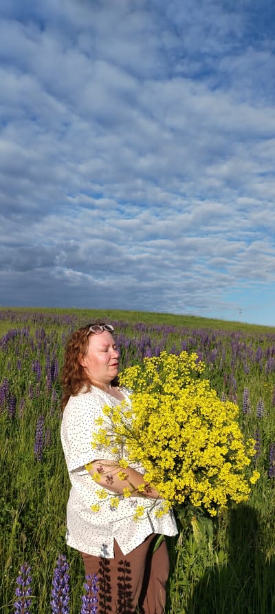 The image, set in Зипуново, Russia, depicts a woman standing in a vibrant green field filled with purple lupines under a partly cloudy blue sky. The woman, positioned slightly left of center, has reddish-brown wavy hair and fair skin. Her eyes are closed, and she has a subtle, serene expression on her face, with red lipstick visible. She wears a white, short-sleeved blouse with black polka dots and brown trousers. White-framed sunglasses are perched on top of her head. She holds a large, dense bouquet of numerous small, bright yellow flowers against her torso, cradled in her arms. Sunlight from the right casts clear shadows from the flowers onto her blouse and trousers, and from her figure onto the tall grass and prominent purple lupines in the foreground. In the background, a gently sloping green hill, also dotted with purple lupines, extends towards the horizon. The sky occupies the upper two-thirds of the image, featuring numerous scattered cumulus clouds, some appearing brighter and others darker, especially on the far left. A distant utility pole is visible on the far right horizon against the green hillside.