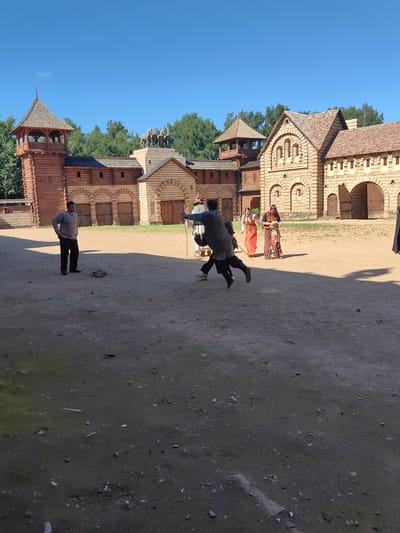A mid-shot captured outdoors on a sunny day shows a historical reenactment or film set. The background features a fortified wooden structure with towers and stone buildings, reminiscent of a medieval settlement. In the foreground, several individuals are present. Two men are engaged in a sword fight, one in chainmail is mid-lunge. Another man in a light-colored shirt and dark pants stands watching the fight. Two women in traditional historical clothing are also present, observing the scene. The ground is dirt and sand, with scattered debris. The sky is clear and blue. The location is identified as Терехово-2 Снт, Russia.