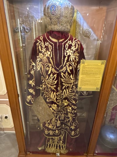 This is a medium shot, interior photograph of religious vestments displayed in a glass-covered wooden case at the Bachkovo Monastery in Bachkovo, Bulgaria. The primary subject is a dark red velvet robe, heavily embroidered with gold thread in a floral and vine pattern. A large, ornate silver crown with a small religious icon at its front sits atop the robe. To the left of the robe, a silver bishop's staff with a serpent design and a cross at the top is visible. A small, gold-embossed book is placed in front of the robe. A yellow informational plaque with text is attached to the glass in front of the robe. The background of the case is dark, with reflections on the glass. The environment outside the case is dimly lit, with parts of the monastery's interior visible, including a wall with a light switch and some decorative elements.