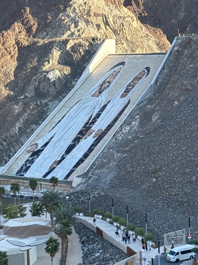 This image depicts a panoramic view of the Hatta Dam in Hatta, United Arab Emirates, showcasing a large mural and surrounding recreational facilities. The prominent feature is a vast mural painted directly onto the inclined, light-colored concrete face of the dam, depicting two male figures dressed in traditional white Emirati thobes and ghutras. Both figures have visible beards and appear to be smiling. Their hands are also visible as part of the artwork. Water cascades from multiple points along the lower edge of the dam, creating small waterfalls into a basin below.
The dam is situated within a rugged mountainous landscape, characterized by steep, rocky, arid slopes displaying shades of brown and grey, particularly evident in the upper left and right portions of the image. Sunlight illuminates parts of the mountains and the dam face, creating areas of shadow and light.
Below the dam, a developed area features a winding, light-colored paved pathway. Numerous individuals, appearing as adults, are walking along this pathway in small groups or alone. The area is landscaped with abundant palm trees and planters containing green shrubbery. Modern, light-colored buildings with distinct architectural designs are visible to the left of the pathway. Dark-colored streetlights are spaced along the route. Towards the bottom right, a white van is parked near a sign structure. The ground immediately surrounding the pathway and beneath the dam is covered in grey and dark gravel or loose rocks.