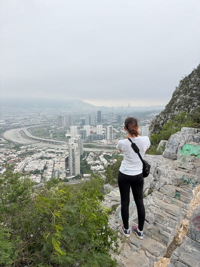 A medium shot shows a woman wearing a white t-shirt and black leggings standing on a rocky cliff edge, leaning forward to look out over the city of Santa Catarina, Mexico. The woman is facing away from the camera, and a black strap is visible across her back. The landscape below is hazy, with buildings and roads stretching into the distance. The cliff edge is composed of gray rocks and sparse vegetation.
